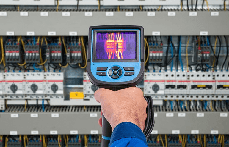 Endüstriyel bir elektrik panosu önünde, elinde termal kamera veya multimetre bulunan, tam donanımlı (KKD'li) bir elektrik mühendisinin ölçüm yaparken çekilmiş yakın plan fotoğrafı.